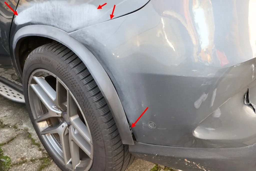 Delle und Kratzer am hinteren Radlauf und Stoßfänger eines grauen SUV, mit Pfeilen markiert